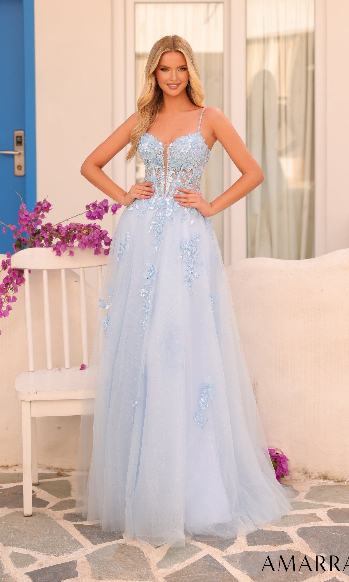 A woman in the Amarra 88086 long formal dress, a light blue ball gown with floral embellishments, lace embroidery, and thin straps, stands outdoors beside a white chair and purple flowers.